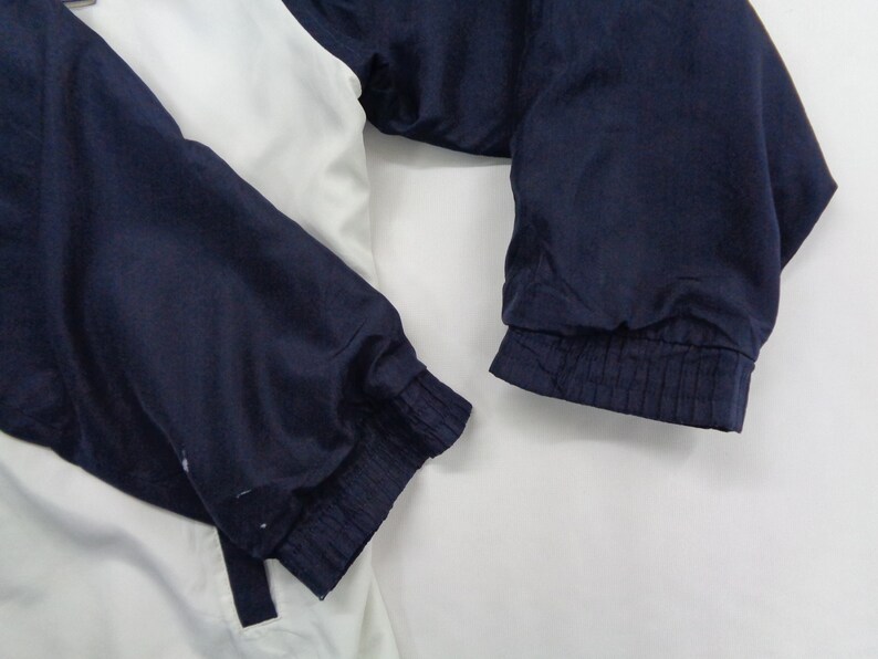 May include: A white and navy blue jacket with elastic cuffs. The jacket has a color block design with white on the upper body and navy blue on the sleeves. The sleeves are gathered at the wrist with elastic.