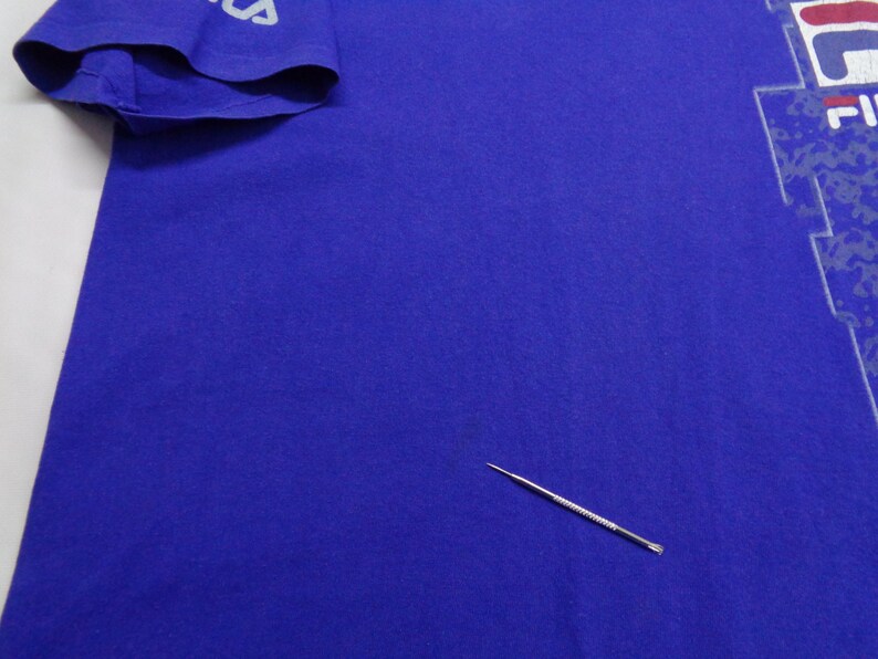 May include: A royal blue t-shirt with a white and red graphic design on the front. The sleeve has the word "FILA" printed in white. The shirt is made of a soft material.