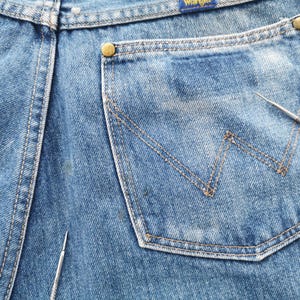 May include: Close-up of blue denim jeans with a Wrangler label. The jeans feature a back pocket with a signature W-shaped stitching design. The pocket is outlined with tan stitching, and the jeans have brass-colored rivets.