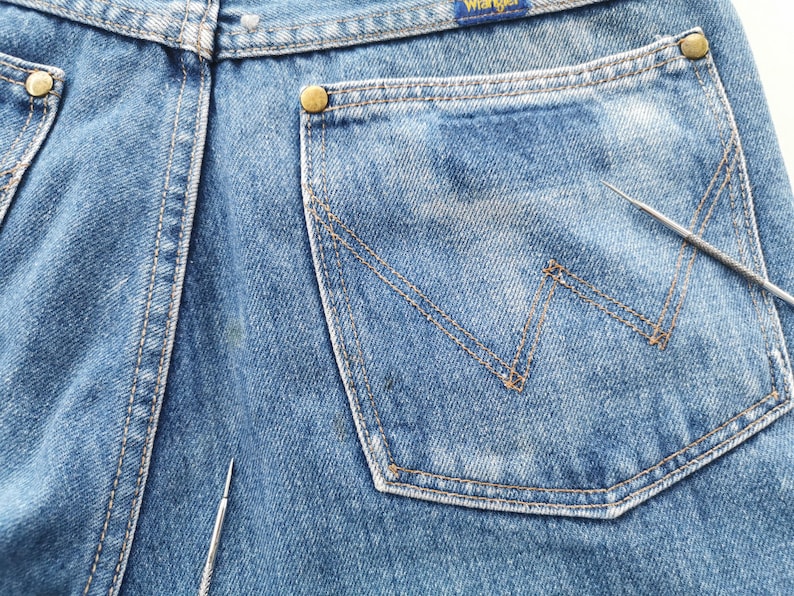 May include: Close-up of blue denim jeans with a Wrangler label. The jeans feature a back pocket with a signature W-shaped stitching design. The pocket is outlined with tan stitching, and the jeans have brass-colored rivets.