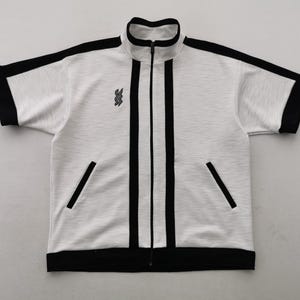 May include: White and grey short-sleeved track jacket with black detailing. It has a full-length zip, black stripes down the front, and black trim on the sleeves and hem. Includes two front pockets and a small emblem on the chest.