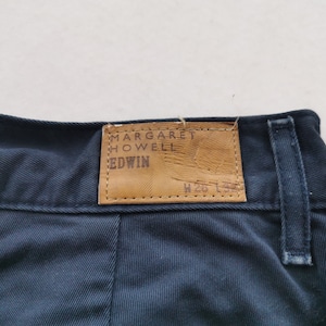 May include: Close-up of navy blue pants with a leather tag. The tag reads "MARGARET HOWELL EDWIN" in embossed lettering. The pants have a classic design with a visible seam and belt loop.