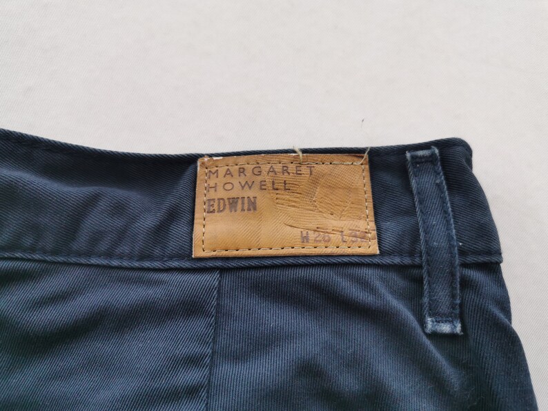 May include: Close-up of navy blue pants with a leather tag. The tag reads "MARGARET HOWELL EDWIN" in embossed lettering. The pants have a classic design with a visible seam and belt loop.
