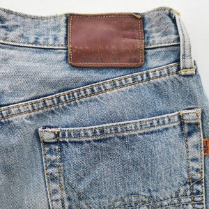 May include: Close-up of light blue denim jeans with a brown leather patch and a back pocket. The jeans have yellow stitching and a small orange tag with the brand name "EDWIN". The leather patch has faded text.