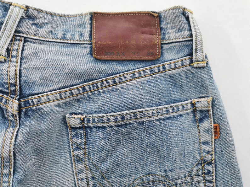 May include: Close-up of light blue denim jeans with a brown leather patch and a back pocket. The jeans have yellow stitching and a small orange tag with the brand name "EDWIN". The leather patch has faded text.