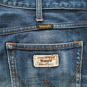 K&ouml;nnte beinhalten: Nahaufnahme der R&uuml;ckseite einer blauen Jeans. Die Jeans hat ein Wrangler-Etikett auf der Ges&auml;&szlig;tasche und am Bund. Die N&auml;hte sind in einer kontrastierenden braunen Farbe gehalten. Die Jeans ist verwaschen und abgenutzt.