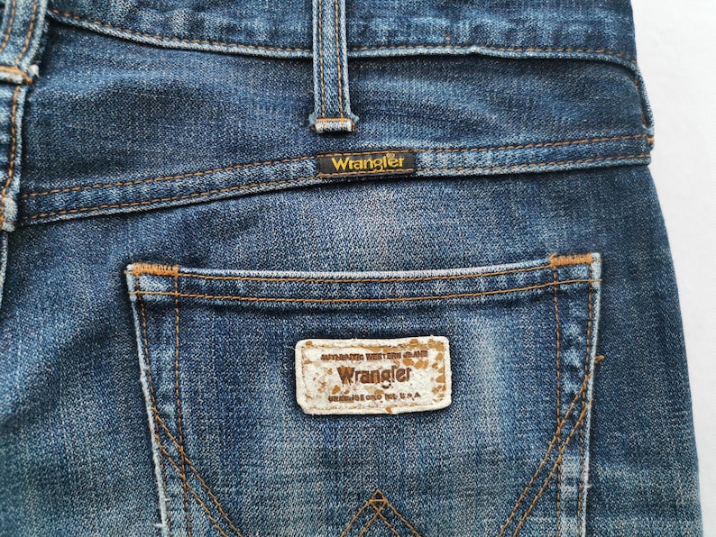 K&ouml;nnte beinhalten: Nahaufnahme der R&uuml;ckseite einer blauen Jeans. Die Jeans hat ein Wrangler-Etikett auf der Ges&auml;&szlig;tasche und am Bund. Die N&auml;hte sind in einer kontrastierenden braunen Farbe gehalten. Die Jeans ist verwaschen und abgenutzt.