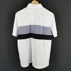 May include: White polo shirt with a color block design. The shirt features a white body with a horizontal stripe in the center composed of gray and black. The shirt has short sleeves and a collared neckline.