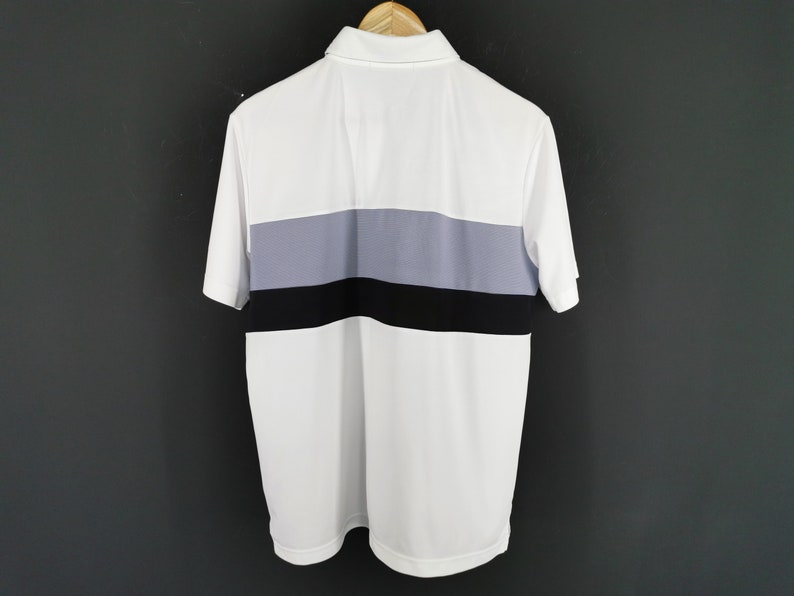 May include: White polo shirt with a color block design. The shirt features a white body with a horizontal stripe in the center composed of gray and black. The shirt has short sleeves and a collared neckline.