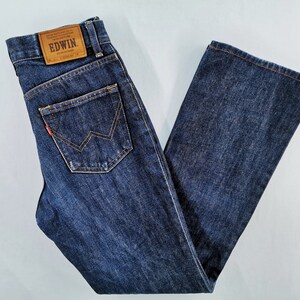 May include: Dark blue denim jeans with a classic five-pocket design. The jeans feature a leather patch with the brand name "EDWIN" and a red logo detail on the back pocket. The jeans are folded neatly, showcasing the straight leg cut.