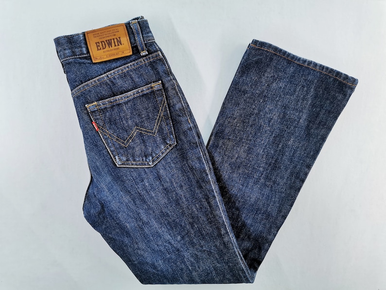 May include: Dark blue denim jeans with a classic five-pocket design. The jeans feature a leather patch with the brand name "EDWIN" and a red logo detail on the back pocket. The jeans are folded neatly, showcasing the straight leg cut.