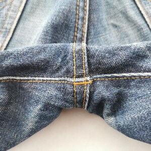 K&ouml;nnte beinhalten: Nahaufnahme einer blauen Jeans, die den Schrittbereich zeigt. Die Jeans hat sichtbare N&auml;hte mit gelben N&auml;hten. Der Stoff hat ein abgenutztes, strukturiertes Aussehen mit helleren und dunkleren blauen Bereichen.