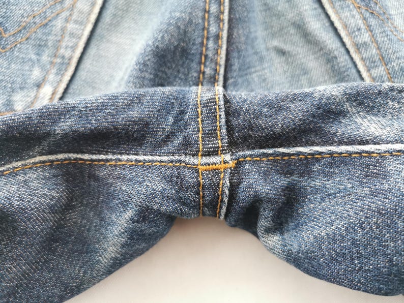K&ouml;nnte beinhalten: Nahaufnahme einer blauen Jeans, die den Schrittbereich zeigt. Die Jeans hat sichtbare N&auml;hte mit gelben N&auml;hten. Der Stoff hat ein abgenutztes, strukturiertes Aussehen mit helleren und dunkleren blauen Bereichen.