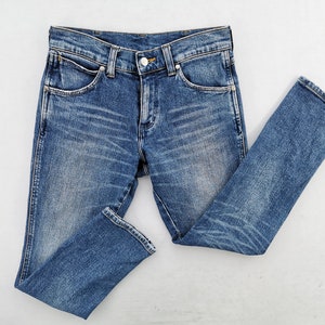May include: Blue denim jeans with a classic five-pocket design. The jeans have a button and zipper closure, belt loops, and a faded wash. The fabric shows subtle wear patterns, and the jeans are laid flat on a white surface.