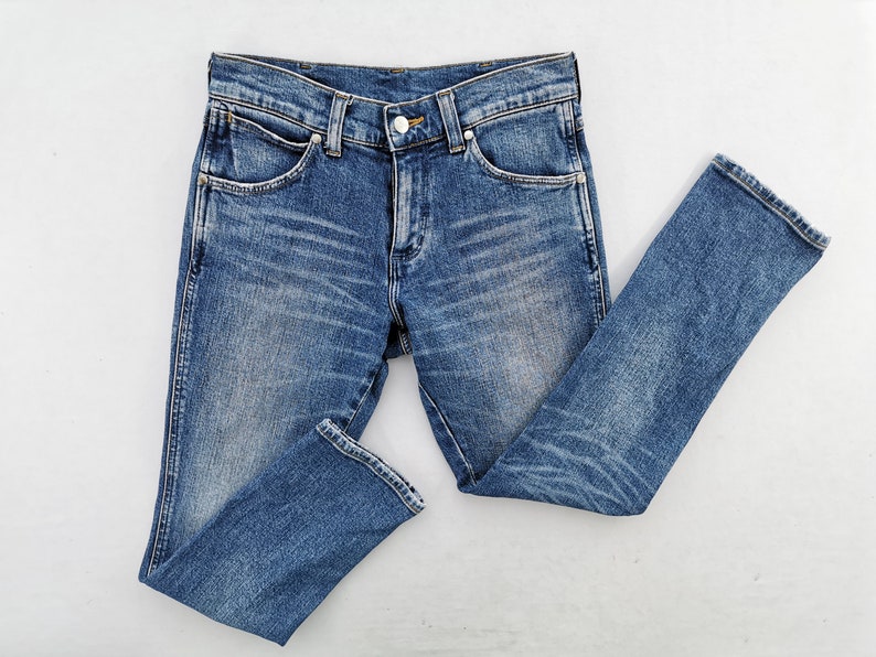 May include: Blue denim jeans with a classic five-pocket design. The jeans have a button and zipper closure, belt loops, and a faded wash. The fabric shows subtle wear patterns, and the jeans are laid flat on a white surface.