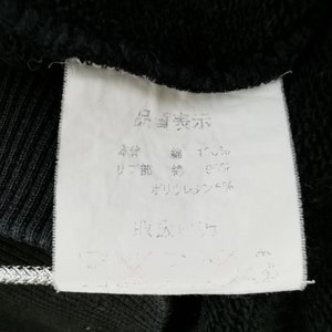 May include: Close-up of a white clothing tag with Japanese text, attached to a black garment. The tag displays text indicating the fabric composition: 100% cotton, 95% cotton rib, and 5% polyurethane.
