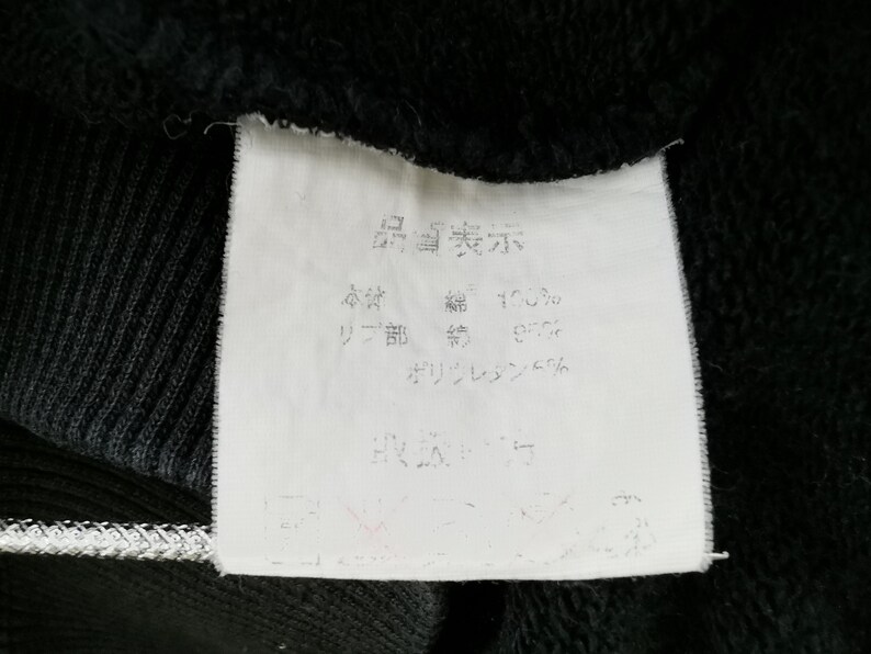 May include: Close-up of a white clothing tag with Japanese text, attached to a black garment. The tag displays text indicating the fabric composition: 100% cotton, 95% cotton rib, and 5% polyurethane.