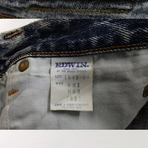 K&ouml;nnte beinhalten: Nahaufnahme einer blauen Jeans mit Acid-Wash-Effekt. Die Jeans zeigt das Markenetikett "EDWIN" und Gr&ouml;&szlig;eninformationen. Die Bundweite ist W81, und die H&uuml;ftweite ist H98 (ca. 81 cm). Es gibt auch ein Pflege- und Fasergehalts-Etikett.