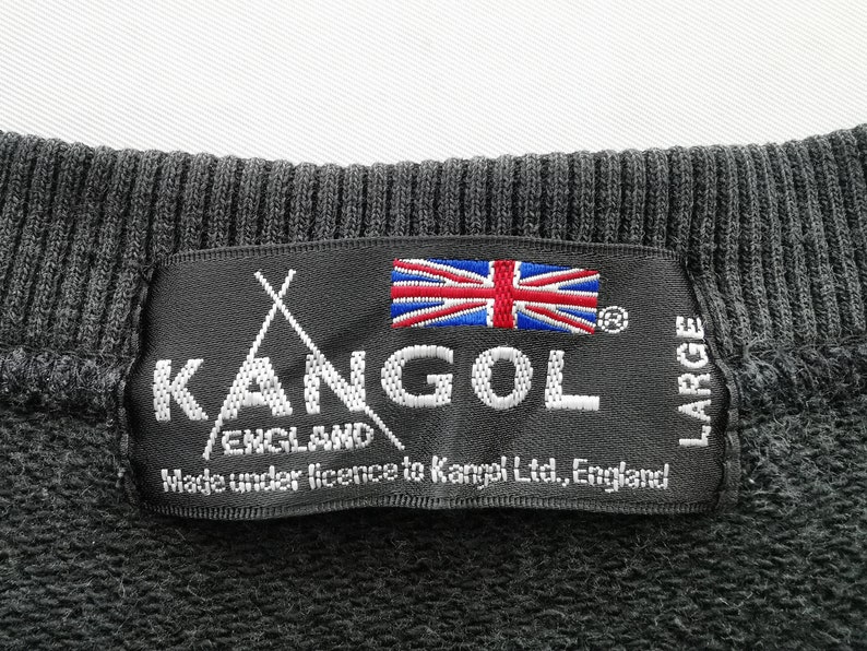 May include: Close-up of a black label from a Kangol England sweatshirt. The label features the Kangol logo, the Union Jack flag, and the text "Made under licence to Kangol Ltd., England." The label also indicates the size is "LARGE."