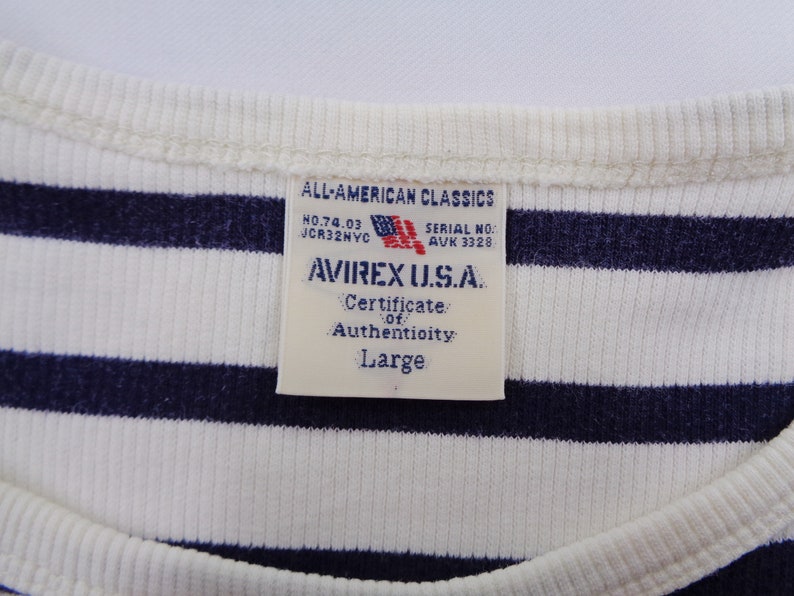 May include: A white and navy blue striped ribbed shirt featuring a rectangular tag at the neckline. The tag reads ALL-AMERICAN CLASSICS, NO.74.03, JCR32NYC, SERIAL NO. AVK 3328, AVIREX U.S.A., Certificate of Authenticity, and the size Large.
