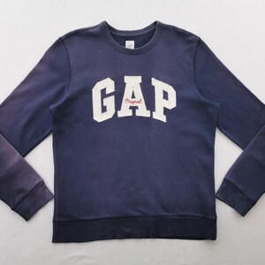 Könnte beinhalten: Ein marineblaues Langarm-Sweatshirt mit Rundhalsausschnitt, das den weißen Schriftzug GAP und das Wort Original in roter Schreibschrift unter dem A zeigt. Das Kleidungsstück hat gerippte Bündchen am Kragen, an den Ärmeln und am Saum.