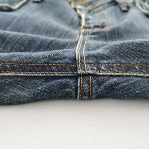 Puede incluir: Primer plano de unos vaqueros de mezclilla azules. La imagen destaca los detalles de las costuras, con hilo amarillo-anaranjado visible. La tela parece desgastada y texturizada, con un enfoque en las costuras y la construcci&oacute;n general de la prenda.