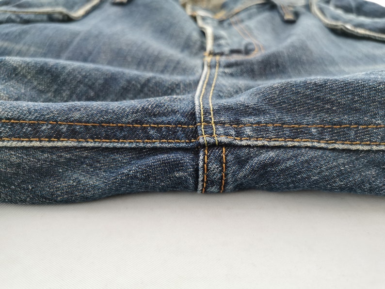 Puede incluir: Primer plano de unos vaqueros de mezclilla azules. La imagen destaca los detalles de las costuras, con hilo amarillo-anaranjado visible. La tela parece desgastada y texturizada, con un enfoque en las costuras y la construcci&oacute;n general de la prenda.