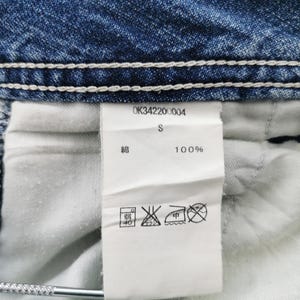 Pu&ograve; includere: Primo piano di un capo in denim con cuciture bianche e un'etichetta bianca. L'etichetta mostra il testo "OK34220004", "S" e "100%" insieme ai simboli di cura. Il denim &egrave; di colore blu medio.