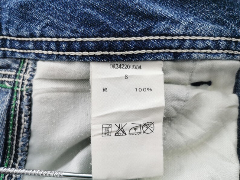 Pu&ograve; includere: Primo piano di un capo in denim con cuciture bianche e un'etichetta bianca. L'etichetta mostra il testo "OK34220004", "S" e "100%" insieme ai simboli di cura. Il denim &egrave; di colore blu medio.