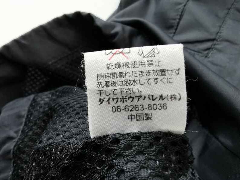 May include: Close-up of a black garment with a white tag. The tag has Japanese text and symbols, including a no-dry-clean symbol. The garment has a mesh lining.