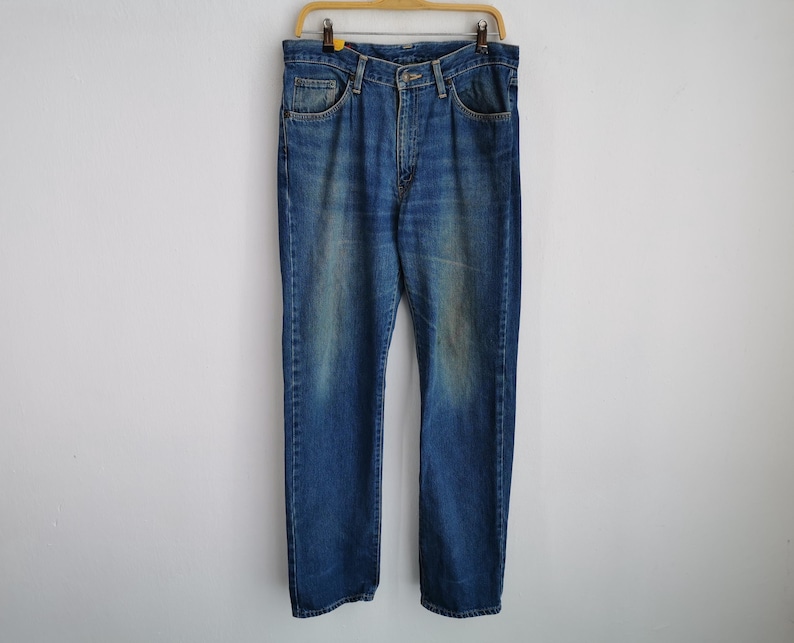 Puede incluir: Vaqueros de mezclilla azul con un lavado descolorido, colgados de una percha de madera. Los vaqueros tienen un dise&ntilde;o cl&aacute;sico de cinco bolsillos y un corte recto. La mezclilla tiene un aspecto desgastado.