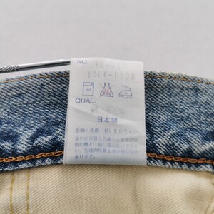 May include: Close-up of a denim garment with a white tag attached. The tag displays text including "NO. KS-010", "1506-0708", and washing instructions. The garment is blue denim with a yellow stitched seam.
