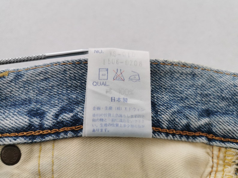 May include: Close-up of a denim garment with a white tag attached. The tag displays text including "NO. KS-010", "1506-0708", and washing instructions. The garment is blue denim with a yellow stitched seam.
