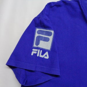 May include: A royal blue t-shirt featuring the FILA logo on the left sleeve. The logo is a white and gray square with the word "FILA" printed below it. The shirt is made of a soft material.