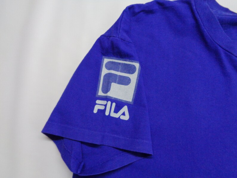 May include: A royal blue t-shirt featuring the FILA logo on the left sleeve. The logo is a white and gray square with the word "FILA" printed below it. The shirt is made of a soft material.