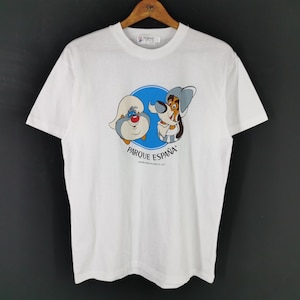 May include: White t-shirt with a cartoon graphic of two characters within a blue circle, and the text "PARQUE ESPAÑA" underneath. The characters are a blue and white bird-like creature and a dog-like character. The shirt is on a wooden hanger.