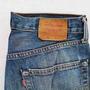 May include: Close-up of blue denim jeans with a brown leather patch. The patch features the Levi Strauss & Co. logo and text. The jeans have a classic five-pocket design and are labeled W 32 L 34.