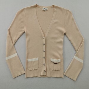 May include: A beige, button-down cardigan with a V-neck and long sleeves. The sweater has two front pockets with white trim and a decorative chain. The sleeves have a white stripe near the cuffs.