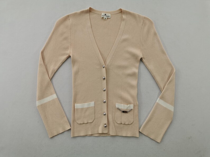 May include: A beige, button-down cardigan with a V-neck and long sleeves. The sweater has two front pockets with white trim and a decorative chain. The sleeves have a white stripe near the cuffs.