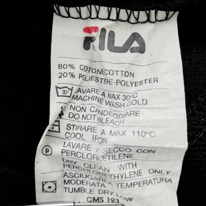 May include: Close-up of a white clothing tag with the FILA logo in red and black. The tag includes washing instructions, fabric composition (80% cotton, 20% polyester), and care symbols. The tag is against a black background.