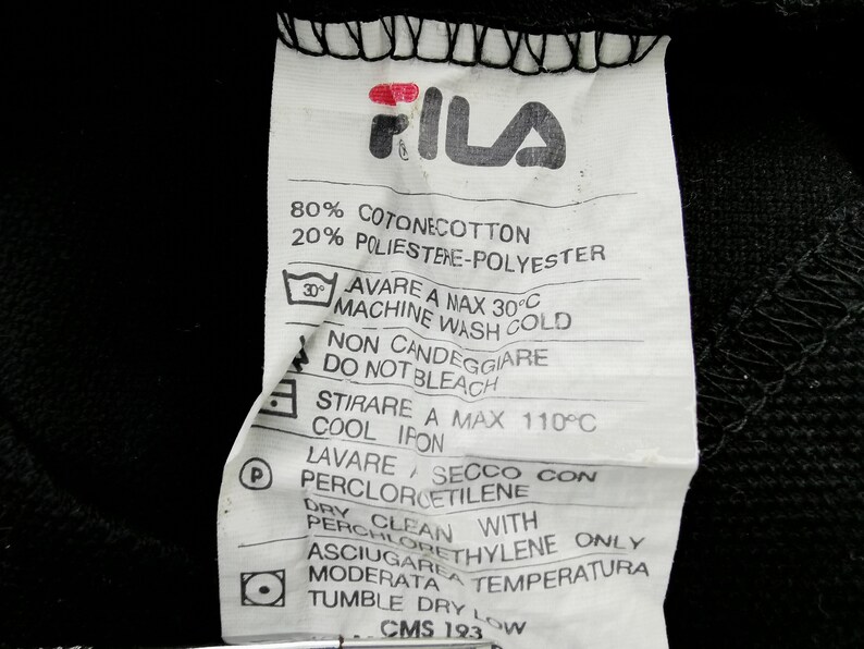 May include: Close-up of a white clothing tag with the FILA logo in red and black. The tag includes washing instructions, fabric composition (80% cotton, 20% polyester), and care symbols. The tag is against a black background.