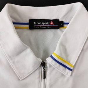 May include: White golf shirt with a zipper closure and a collar. The collar features blue and yellow stripes. The shirt has the "le coq sportif" logo and "GOLF COLLECTION" text on the label.