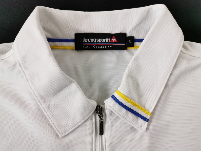 May include: White golf shirt with a zipper closure and a collar. The collar features blue and yellow stripes. The shirt has the "le coq sportif" logo and "GOLF COLLECTION" text on the label.