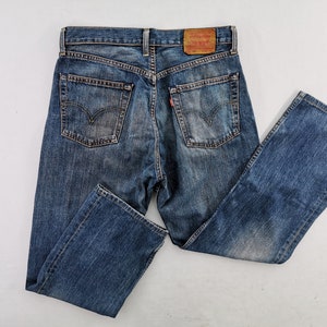 May include: Blue denim jeans with a classic five-pocket design. The jeans feature a faded wash and a leather patch on the back waistband. The back pockets have a signature stitch design.