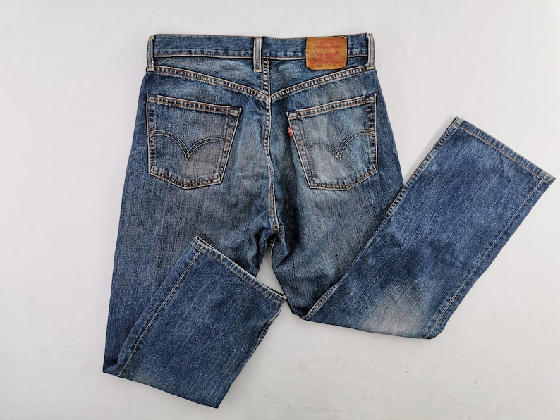 May include: Blue denim jeans with a classic five-pocket design. The jeans feature a faded wash and a leather patch on the back waistband. The back pockets have a signature stitch design.
