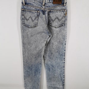 K&ouml;nnte beinhalten: Jeans aus Acid-Wash-Denim mit einem hellblauen und grauen Muster. Die Jeans haben zwei Ges&auml;&szlig;taschen mit einem aufgestickten Design und einen braunen Lederaufn&auml;her mit dem Markennamen "EDWIN". Die Jeans h&auml;ngen an einem Holzb&uuml;gel.