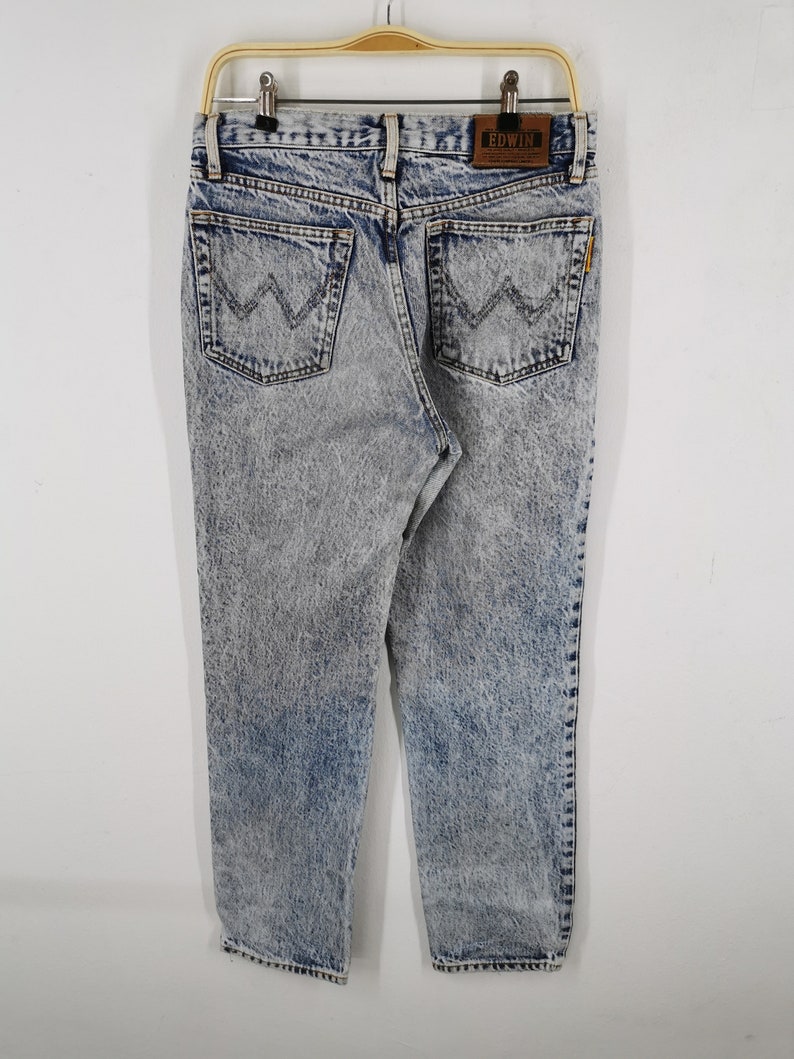 K&ouml;nnte beinhalten: Jeans aus Acid-Wash-Denim mit einem hellblauen und grauen Muster. Die Jeans haben zwei Ges&auml;&szlig;taschen mit einem aufgestickten Design und einen braunen Lederaufn&auml;her mit dem Markennamen "EDWIN". Die Jeans h&auml;ngen an einem Holzb&uuml;gel.