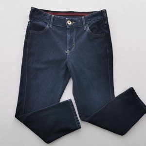 K&ouml;nnte beinhalten: Dunkelblaue Jeans mit klassischem F&uuml;nf-Taschen-Design. Die Jeans haben einen Knopf- und Rei&szlig;verschluss, einen goldfarbenen Knopf und einen geraden Beinverlauf. Die N&auml;hte sind in einer kontrastierenden hellen Farbe gehalten. Die Jeans sind sauber auf einer wei&szlig;en Oberfl&auml;che gefaltet.
