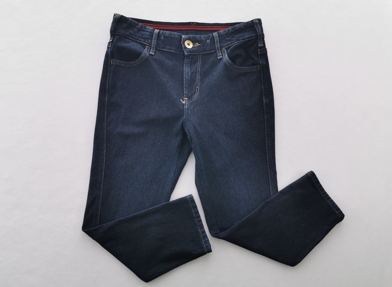 K&ouml;nnte beinhalten: Dunkelblaue Jeans mit klassischem F&uuml;nf-Taschen-Design. Die Jeans haben einen Knopf- und Rei&szlig;verschluss, einen goldfarbenen Knopf und einen geraden Beinverlauf. Die N&auml;hte sind in einer kontrastierenden hellen Farbe gehalten. Die Jeans sind sauber auf einer wei&szlig;en Oberfl&auml;che gefaltet.