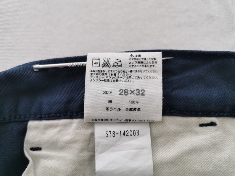 May include: Close-up of navy blue denim pants with a white interior. A tag displays the size 28x32, along with washing instructions and fabric composition. The tag also includes Japanese text and the number 578-142003.